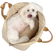 Hvid bomuldshund ligger i en sandfarvet hundetaske med tungen på snuden